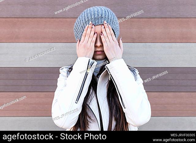 Teenage girl covering her face with hands while standing against wall