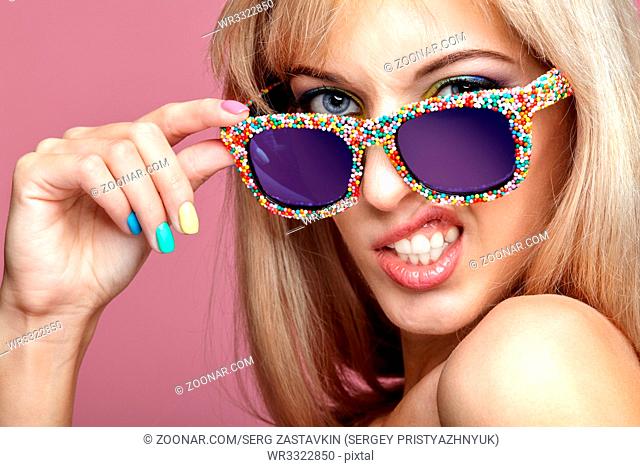 Young blonde woman with fun candy glasses on pink background. Portrait of female in lilac dress with blue and yellow eyes makeup making grimace