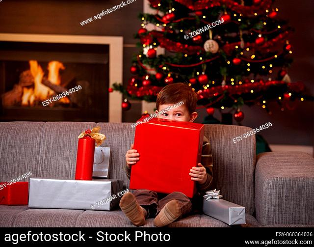 Little boy sitting on couch hiding behind gift box, looking over it