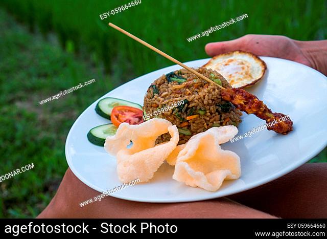 Nasi goreng, traditional Indonesian dish served with prawn crackers and chicken satay on white plate. Closeup woman's hands holding plate with fried rice while...