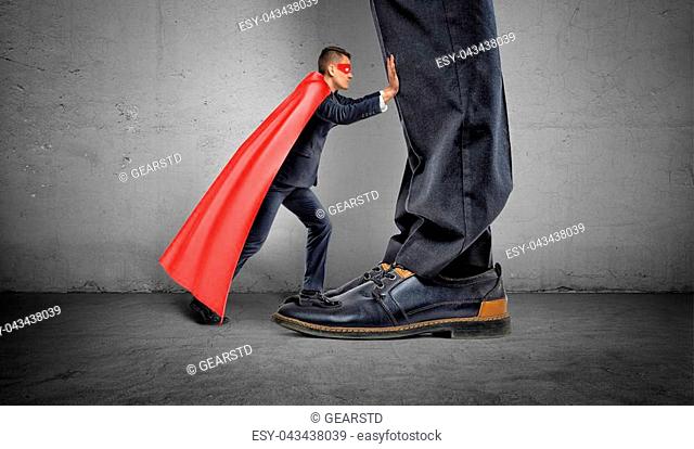 A small businessman in a superhero cape trying to push away giant man legs. Uneven competition. Best performance. Business problems