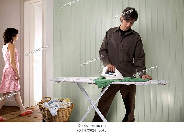 A man ironing whilst a woman is walking into another room