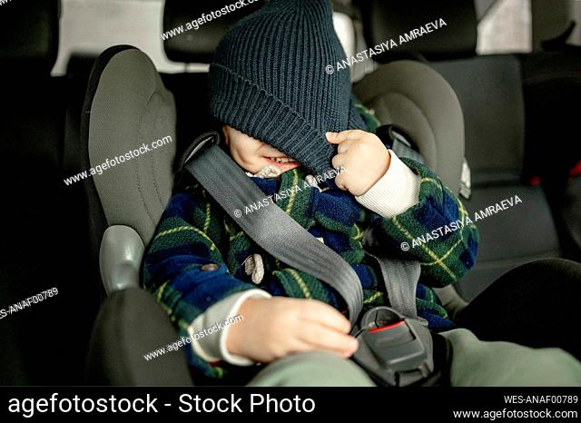 Happy boy covering face with knit hat in car safety seat