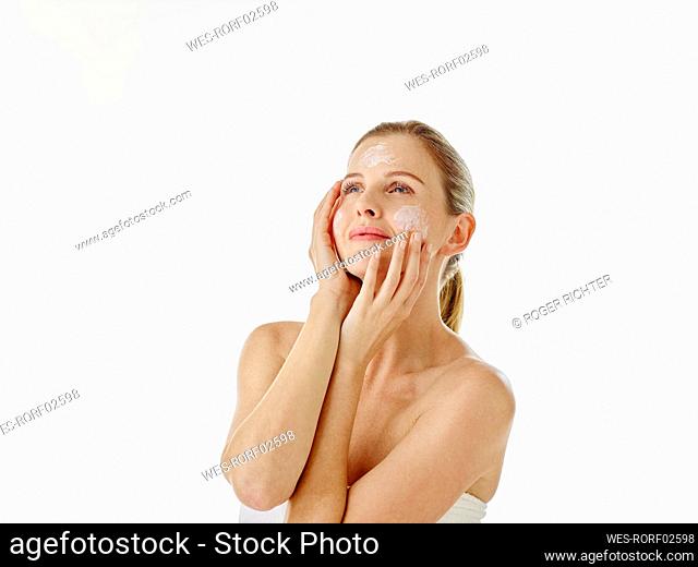 Thoughtful woman wrapped in towel looking away while applying cream standing against white background