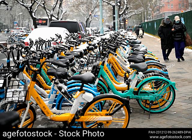 12 December 2023, China, Peking: Two people walk past parked rental bikes, some of which are covered in snow. Many people in Beijing use the rental bikes to get...