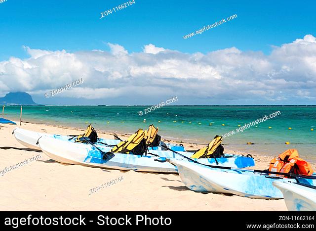 Blue and white kayaks with life jackets on sandy beach at Mauritius island, sunny day with clouds in the sky and mountain background