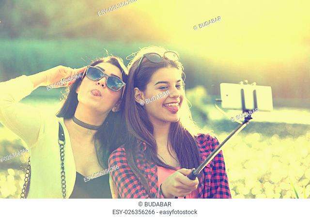 Two pretty teenage girls taking selfie on mobile phone with stick in the nature beside river. Positive and crazy emotions