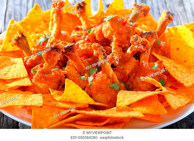close-up of deep fried battered crispy chicken wing lollipops sprinkled with parsley and finely chopped chilli with nachos on a white plate on dark wooden table