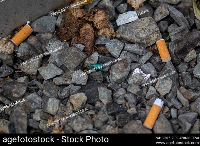 SYMBOL - 03 July 2023, Berlin: Remains of a syringe, a piece of aluminum foil and cigarette butts lie on the ground next to the entrance to public mobile...