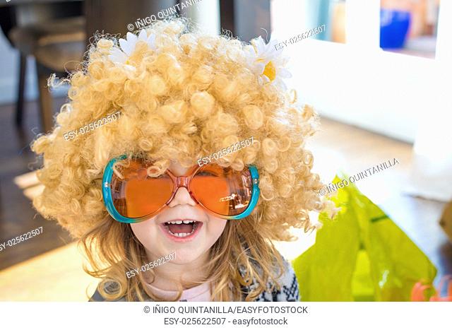 funny portrait of three years old child disguised as sixties, with great curly blond hair wig with daisy flowers on head, with orange and green colorful glasses