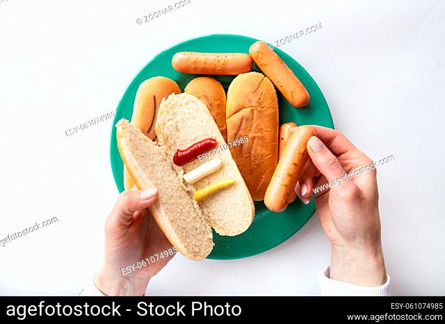 Fresh hot dog buns which lie on a green plate along with sausages. The girl holds in her hands a bun with sauces in which she puts a sausage