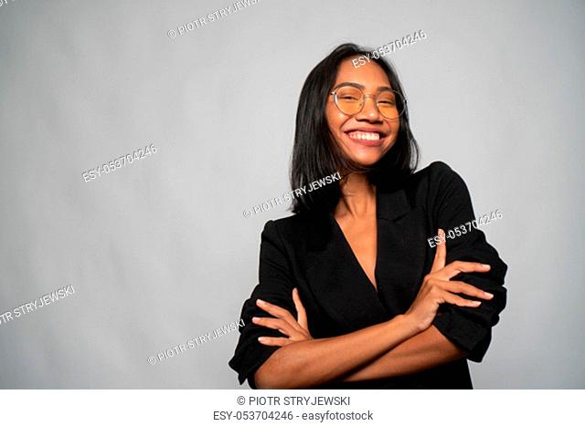 Portrait of attractive young Asian smiling woman in elegant black blazer and glasses posing over light gray wall background. Mock up copy space
