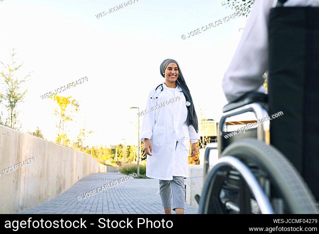Smiling female healthcare worker walking toward patient sitting on wheelchair