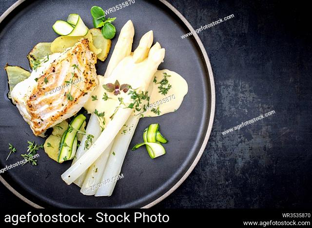 Modern German fried cod fish filet with white asparagus in hollandaise sauce with roast potatoes and sliced zucchini as top view on a plate with copy space...