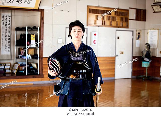 Female Japanese Kendo fighter standing in a gym, holding Kendo mask and sword, looking at camera