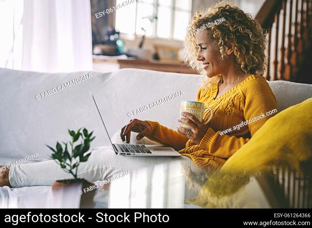 Real life of woman using laptop computer smilig and drinking a coffee at home comfortably sitting on the sofa. Modern female people use computer notebook in...