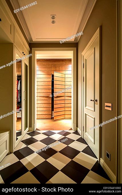 Illuminated modern corridor with light walls and a parquet with chess tiles on the floor. There is a light wardrobe with mirrors