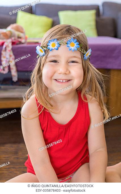portrait of three years old blonde pretty girl face smiling, with red sleeveless t-shirt and diadem with flowers, sitting indoor at home