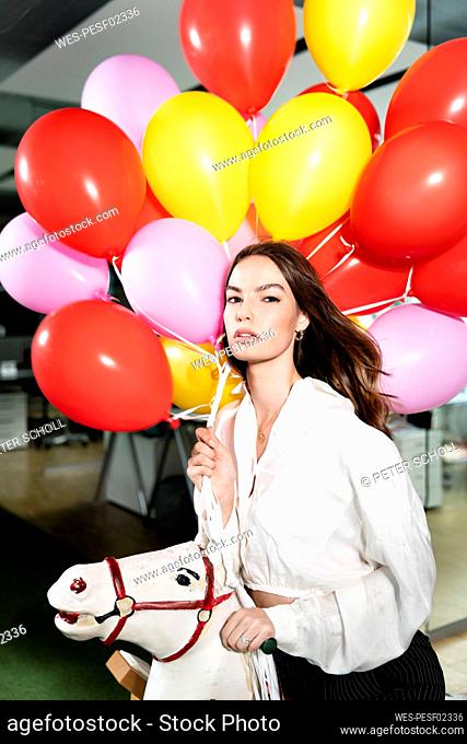 Businesswoman holding colorful balloon while sitting on rocking horse at office