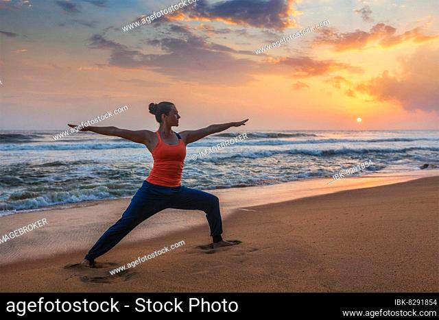 Woman doing Hatha yoga asana Virabhadrasana 1 Warrior Pose outdoors on ocean beach on sunset. Kerala, India, Asia