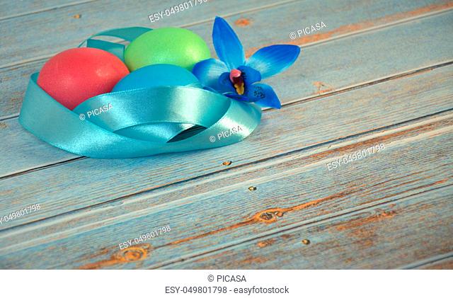 Three Easter eggs with a blue satin ribbon and an orchid bud on a wooden table. Close-up