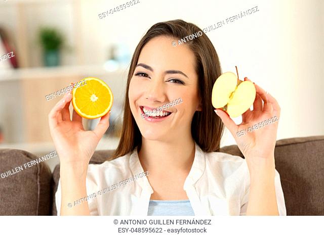 Front view of a happy woman showing half apple and half orange sitting on a couch in the living room at home