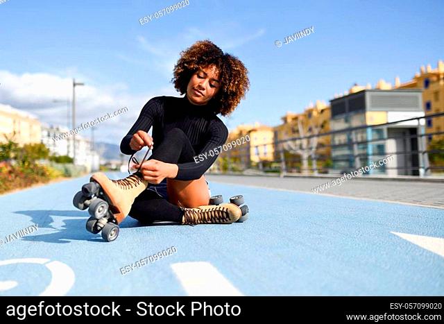 Young smiling black girl sitting on bike line and puts on skates. Woman with afro hairstyle rollerblading on sunny day