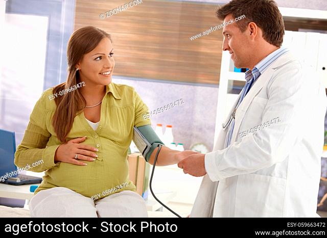 Smiling pregnant woman sitting in consulting room, doctor checking blood pressure