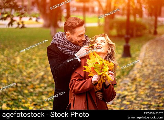 Happy young people in love, man hugging woman from behind stroking her cheek, happy couple walking in a autumn park wearing stylish coats and picking up fallen...