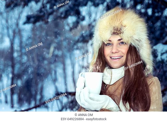 Portrait of a beautiful young girl in a stylish fur hat standing in a snowy park and warming hands on a hot mug of tea, enjoying the beautiful winter view of...