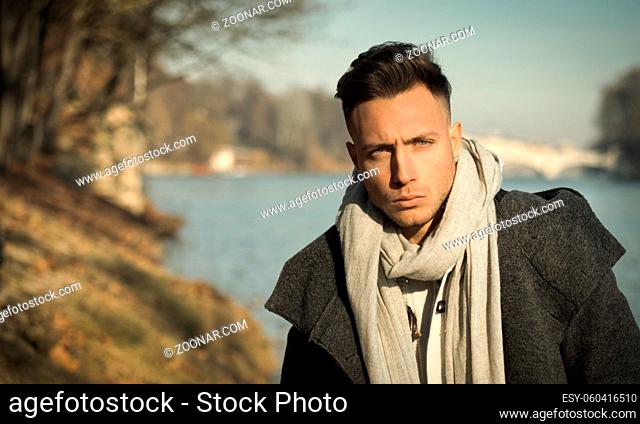 One handsome young man in urban setting in European city, Turin in Italy by the river Po, in cold winter day, looking at camera