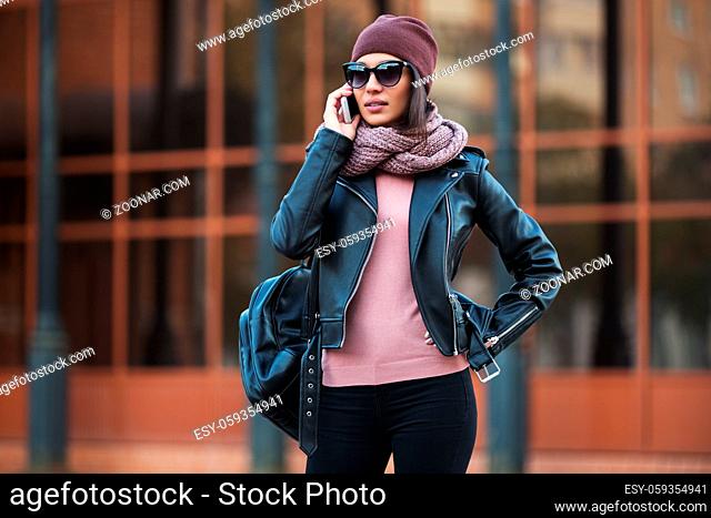 Young fashion woman in black leather jacket using cell phone in city street. Stylish female model wearing sunglasses beanie and scarf outdoor