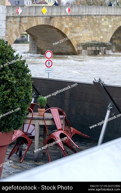 21 November 2023, Bavaria, Regensburg: Chairs and a table stand behind a flood protection wall on the bank of the flooding Danube near the Stone Bridge