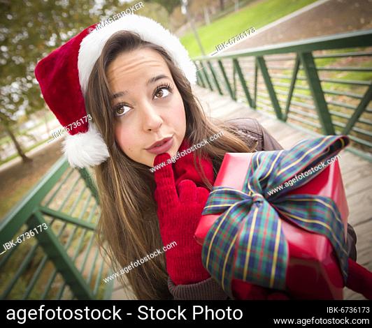 Pretty festive smiling woman wearing a christmas santa hat with wrapped gift and bow outside
