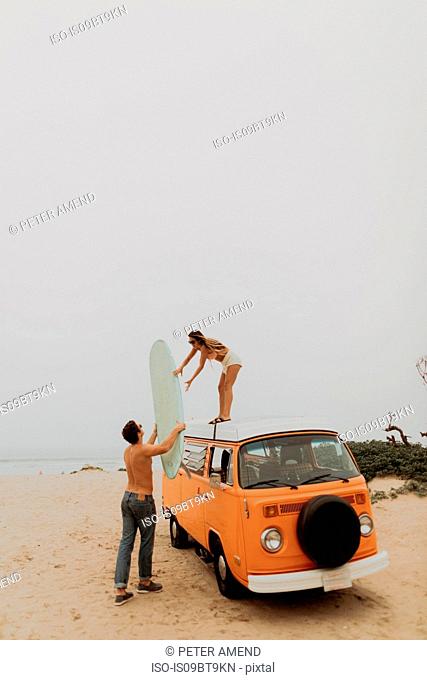 Young surfing couple unloading surfboard from top of recreational vehicle on beach, Jalama, Ventura, California, USA