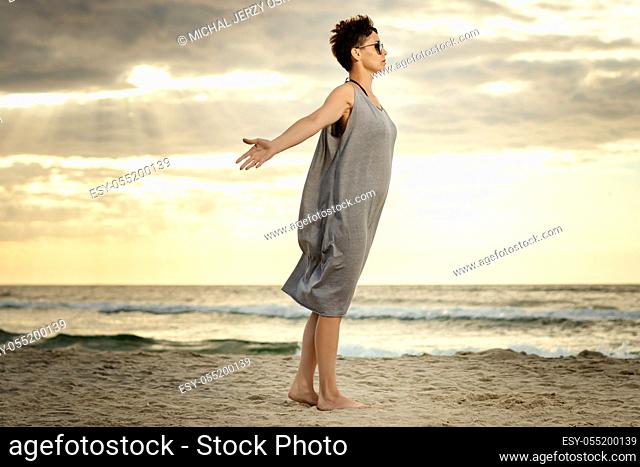 beautiful woman in a gray dress spreads her hands on the beach at sunset