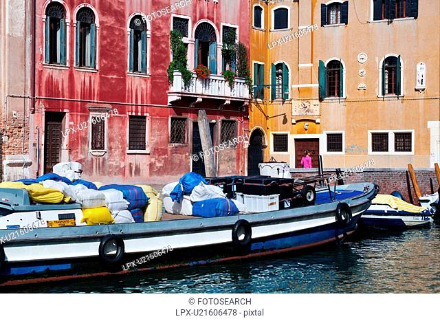 Bright blue and white laden barge moored in front of red and yellow palazzi, with single woman in pink walking alongside canal, sunny autumn morning in Venice