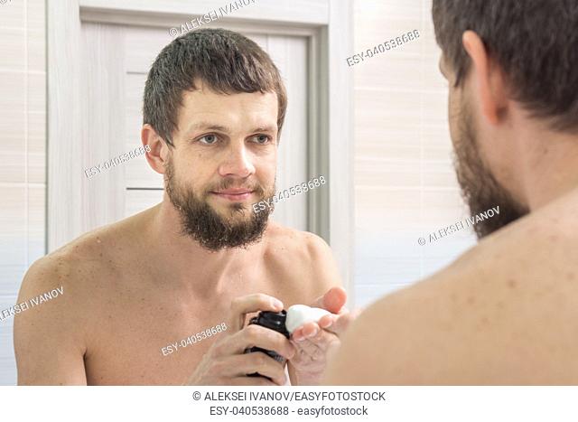 An unshaven man applies a shaving foam on the palm of his hand, and looks in the mirror in the bathroom
