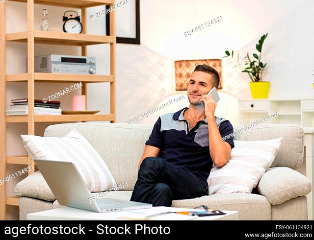 Smiling man taking mobile or smart phone and starting talking over it. Picture of handsome young man sitting on sofa at home and looking in front of him