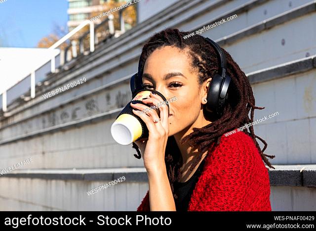 Young woman listening music while drinking coffee from reusable cup on sunny day