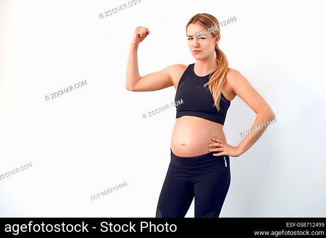 Fun loving young pregnant woman with bare baby belly standing flexing her arm to show her muscles with a wry smile over a white studio background
