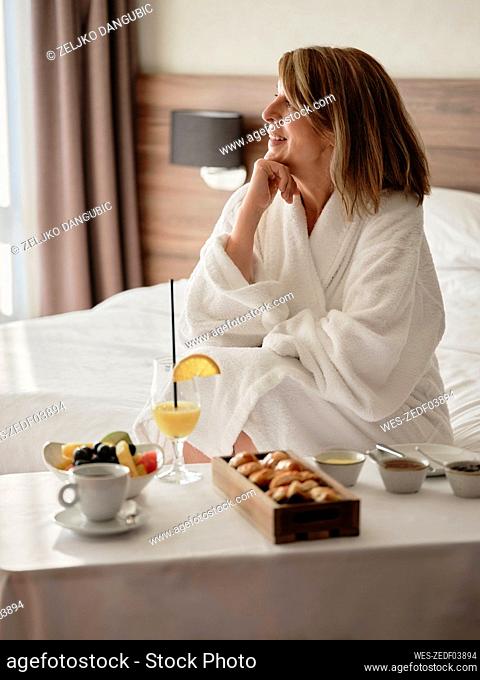 Smiling thoughtful senior woman looking away while sitting with breakfast in luxury hotel room