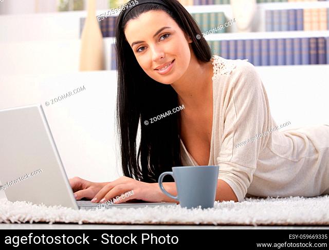 Attractive woman lying on carpet using laptop computer at home, having coffee, smiling at camera