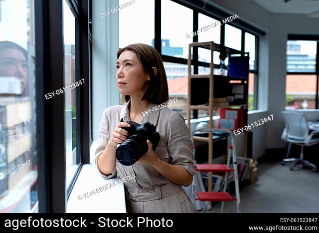 Asian businesswoman holding camera looking through window in creative office