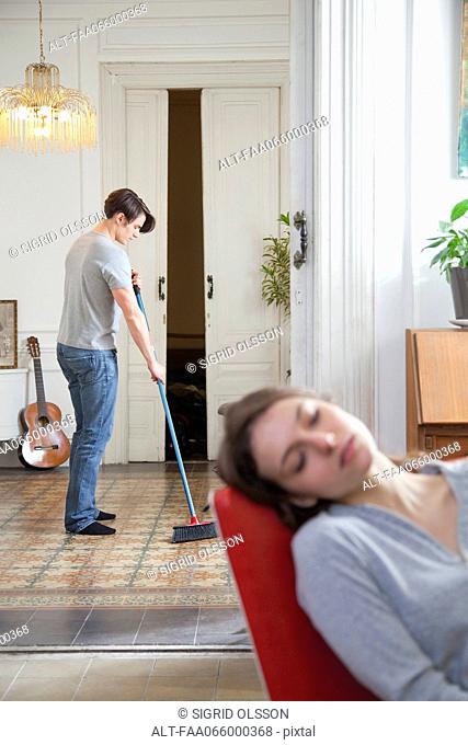 Man sweeping floor at home, woman napping in foreground