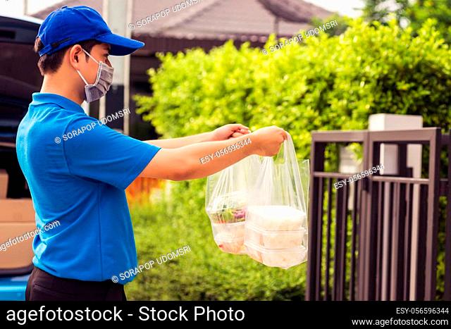 Asian young delivery man in blue uniform wearing face mask making grocery service giving rice food boxes plastic bags and coffee at front house under pandemic...