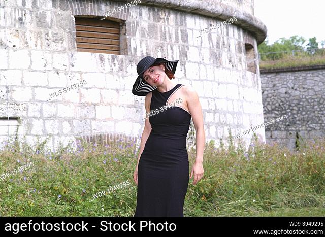 young woman in form-fitting black dress, with happy face, portrayed against fortress walls
