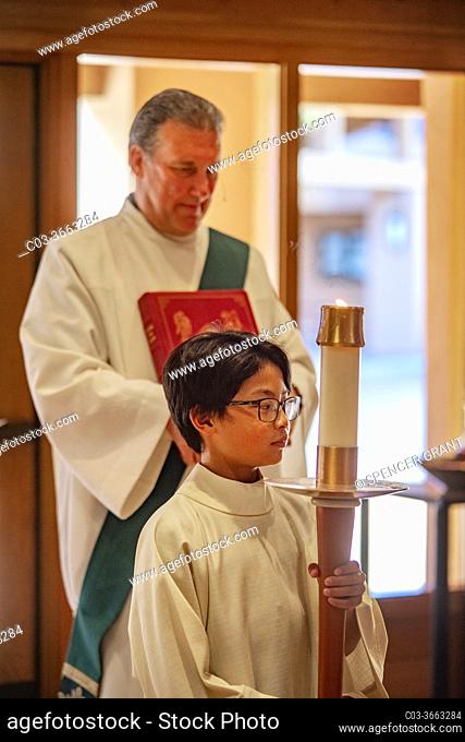 A young robed boy altar assistant holds a ceremonial candle while waiting to participate in a procession to mass along with a book carrying deacon during mass