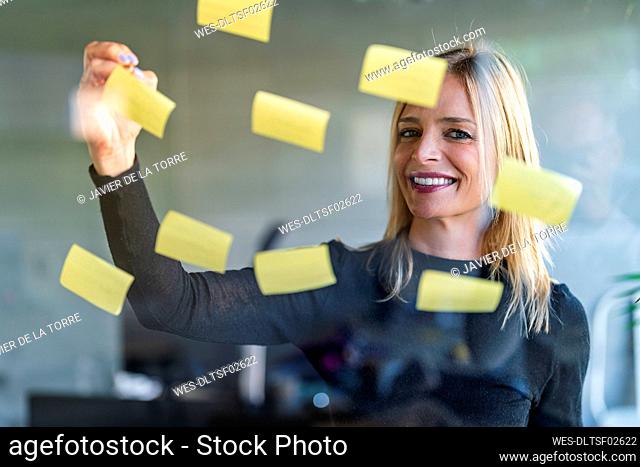 Smiling businesswoman writing on adhesive notes seen through glass wall at office