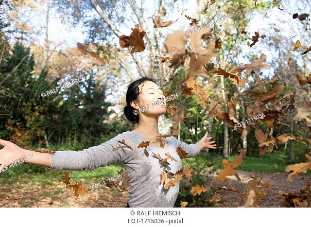 Dry leaves falling on happy woman standing with arms outstretched at park during autumn